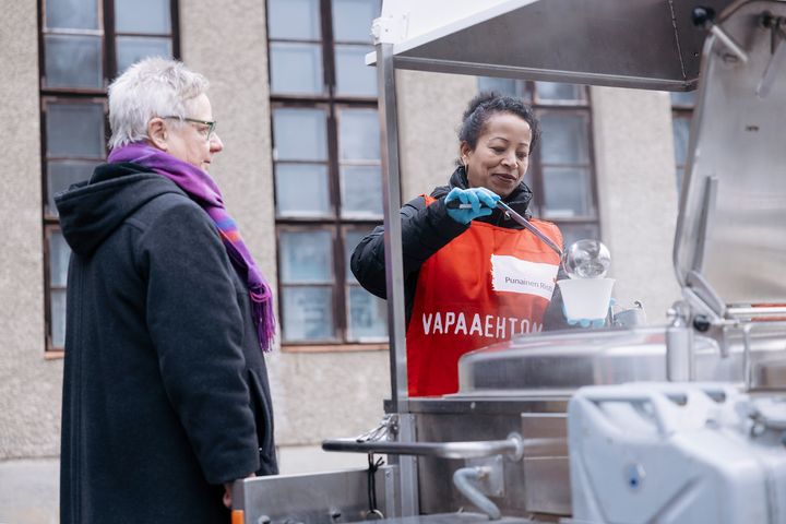 Kaksi henkilöä ruokajakelupisteellä. Vapaaehtoinen jakaa ruokaa.