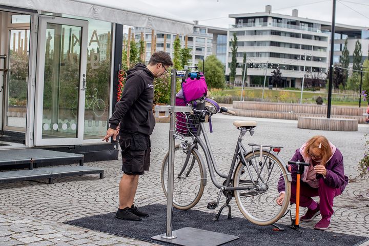 Henkilö pumppaa ilmaa polkupyörän renkaaseen pyöräkeskuksessa ulkona. Toinen henkilö seisoo vieressä.