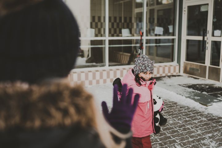 Kuvan etualalla vanhempi vilkuttaa lapselle, joka on nalle sylissä eskarin ulko-ovilla menossa sisään. Lapsi hymyilee ja vilkuttaa takaisin.