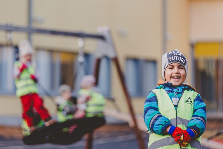 Lapsi nauraa ulkona leikkipaikalla, taustalla muita lapsia keinuilla.