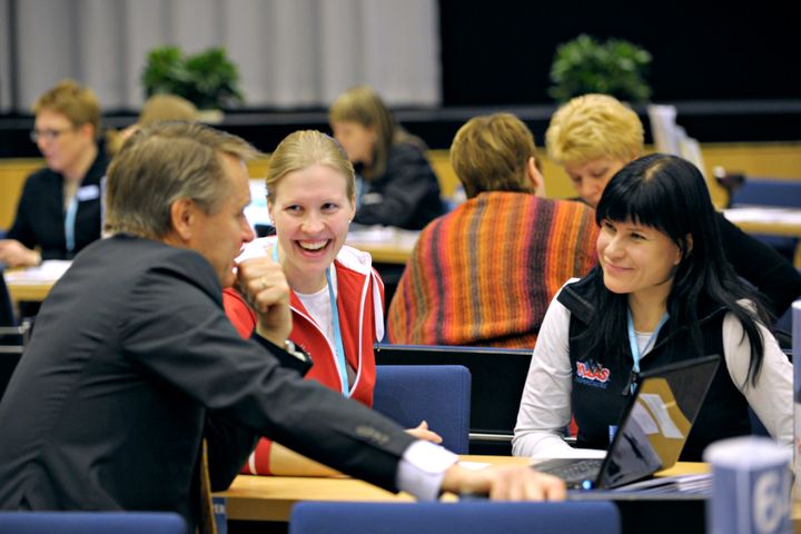 Kolme henkilöä keskustelee iloisesti pöydän ääressä konferenssitilassa.