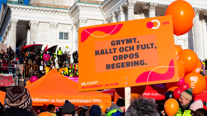 Orange skylt med texten JHL. Bakom skylten syns ett soligt Senatstorg, Helsingfors domkyrka och ljusa moln på himlen.