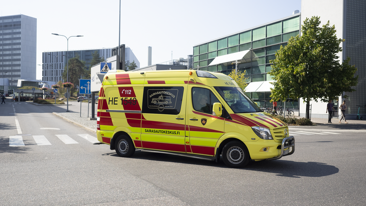 Rödgul ambulans svänger i en korsning i solsken, i bakgrunden byggnader på sjukhusområdet.