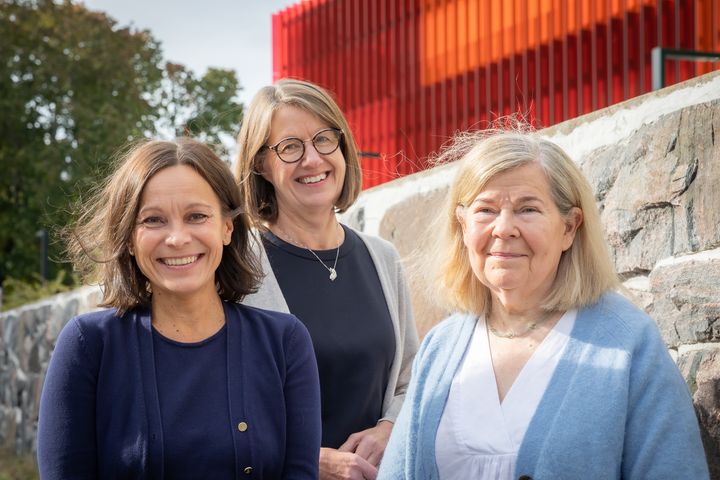 Tre leende kvinnor står utomhus framför en stenmur med ett rött byggnad i bakgrunden.