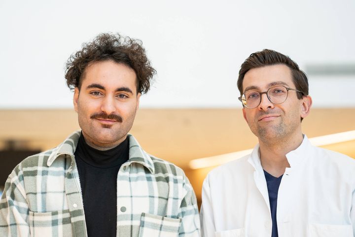 Two people standing side by side, one wearing a plaid shirt over a black turtleneck, and the other in a white button-up shirt with glasses.