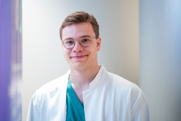 A person wearing glasses and a white lab coat, smiling in a well-lit room.