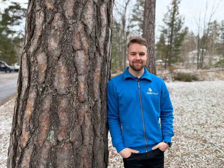 Mies sinisessä takissa seisoo ulkona puun vieressä. Taustalla talvinen metsämaisema.