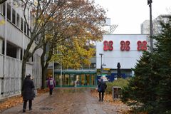 Framför varuhuset går människor på en gata täckt av orange blad. Framför byggnaden står ett träd som fäller löv och i högra kanten två granar.