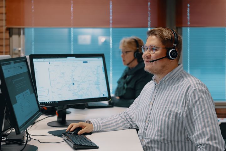 Person med headset som sitter framför två datorskärmar och arbetar. En annan person med headset sitter i bakgrunden.