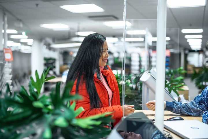 A woman in a red jacket stands at a counter at customer service, interacting with another person.