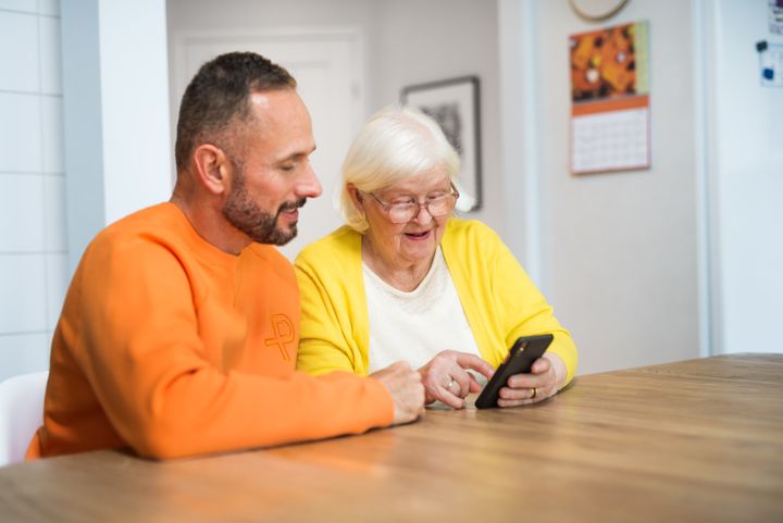 En äldre kvinna och en man sitter vid ett bord och tittar på en mobiltelefon tillsammans.