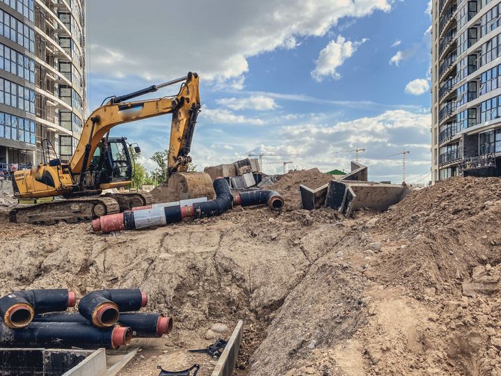 Kaivinkone työskentelee rakennustyömaalla kahden korkean rakennuksen välissä. Maassa on putkia ja rakennusmateriaaleja.