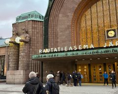 Alt text: Människor går in vid Helsingfors järnvägsstation, där ljusskylten för "RAUTATIEASEMA" är synlig.