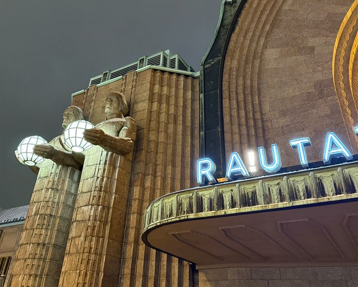 View of a building with statues holding spherical lights and partially visible neon sign reading "RA!UTATIEASEMA".