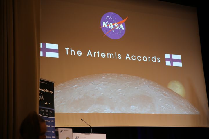 NASA presentation slide with the title "The Artemis Accords" and images of the moon and planets. Finnish flags are displayed. A banner is partially visible in the foreground.