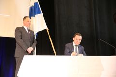 Minister signing a document at a table, with a person standing beside him and a flag in the background.