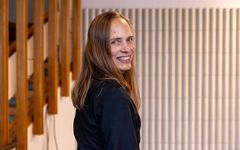 En tiedä, kuka kuvassa on, mutta nainen seisoo sisätiloissa hymyillen kameraan. Taustalla on puukaiteita ja valkoisia pystylaattoja.