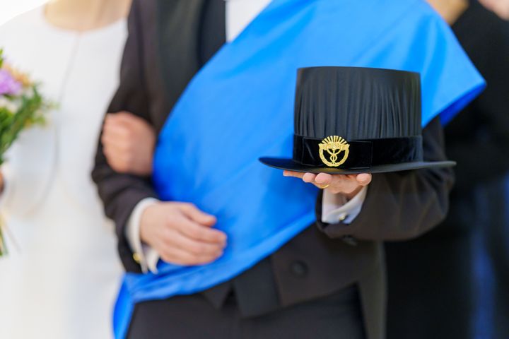 Henkilö kantaa mustaa tohtorinhattua ja sinistä nauhaa.