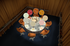 Ympyrän muotoinen pöytä, jonka ympärillä on kahdeksan oranssia tuolia. Pöydän keskellä on kakku ja erilaisia juomia. Pöydän yläpuolella on leijuvia nimilappuja. Taustalla on keltaiset verhot.