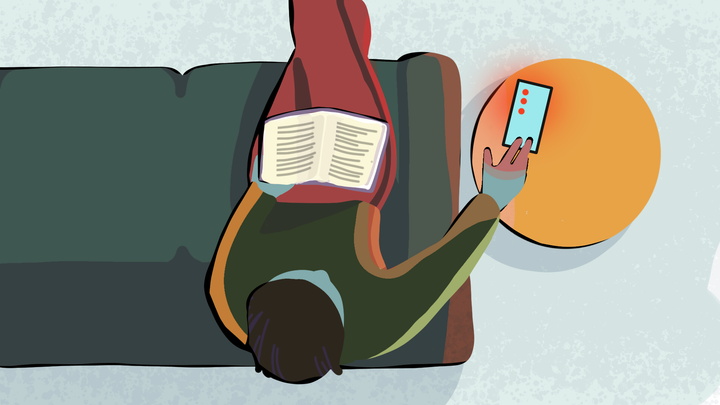 Henkilö istuu sohvalla, lukee kirjaa ja samalla katsoo älypuhelinta.