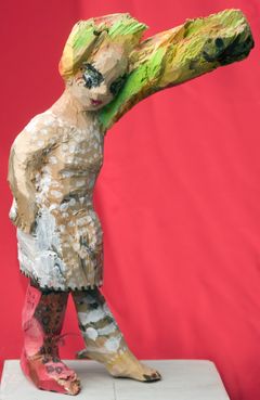 A colorful, abstract sculpture resembling a superhero figure with a raised arm, set against a red background.