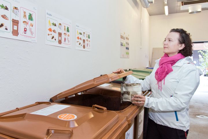 Nainen avaa ruskean roskasäiliön kantta jätehuoneessa ja pitää kiinni biojäteastiasta.