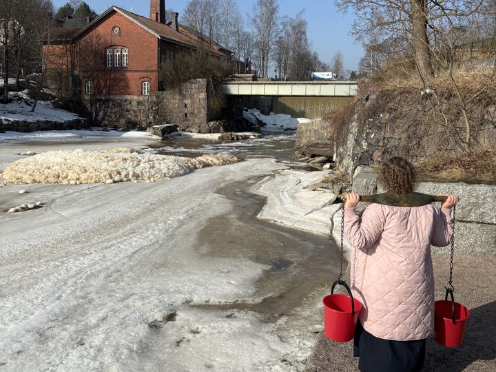 Nainen kantaa ämmänlänkiä, jossa on kaksi punaista ämpäriä. Taustalla näkyy Vantaanjoki ja tiilirakennus.
