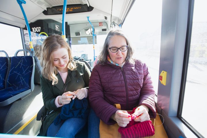 Kuvassa kaksi ihmistä istuu vierekkäin bussissa. Taustalla olevilla penkeillä on tyhjää tilaa.