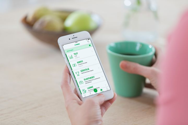 Henkilö pitää kädessään älypuhelinta, jossa on avoinna S-Pankin mobiilisovellus. Sovelluksen etusivulla näkyvät eri osiot, kuten tilit, kortit, säästäjä, sijoitukset ja lainat. Toisessa kädessä on vihreä kahvimuki, ja taustalla näkyy pöytä, jolla on hedelmiä.