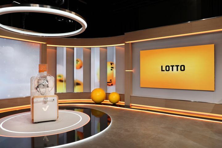 "Lottostudion sisustus: oransseja ja harmaita sävyjä, näkyvillä on iso näyttö, jossa lukee 'Lotto', ja arvontakone."