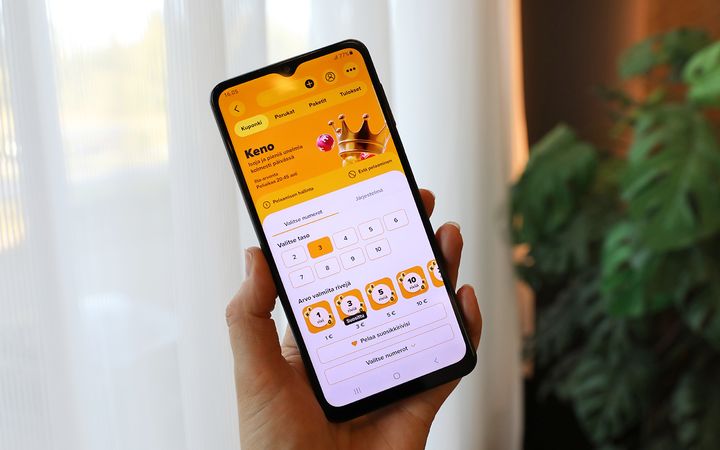 Käsi pitelee älypuhelinta, jonka näytöllä on keltaoranssisävyinen Veikkauksen Keno-pelivalikko; taustalla näkyy vaalea verho ja vihreä huonekasvi.