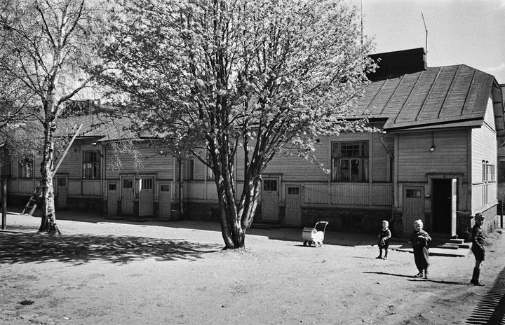 Mustavalkoisessa kuvassa puutalo, lapsia ja puita.