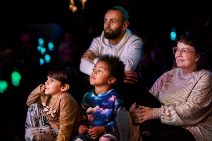 Kaksi lasta ja kaksi aikuista seuraa esitystä salissa, jonka valot on himmennetty.