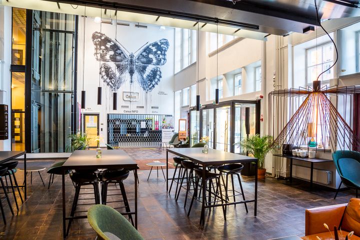 Valoisa ja moderni aula, jossa suurikokoinen mustavalkoinen perhosmaalaus seinällä. Tilassa on pöytiä, tuoleja ja kasveja.