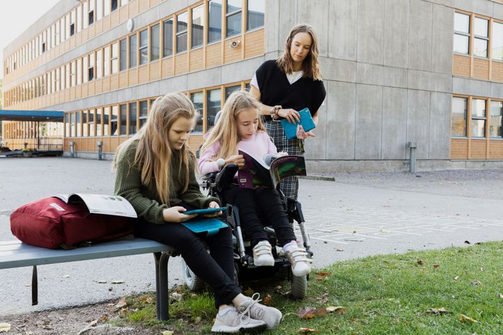 Kaksi koululaista tekee koulun pihalla koulutehtäviä avustajan mahdollistaessa taustalla.