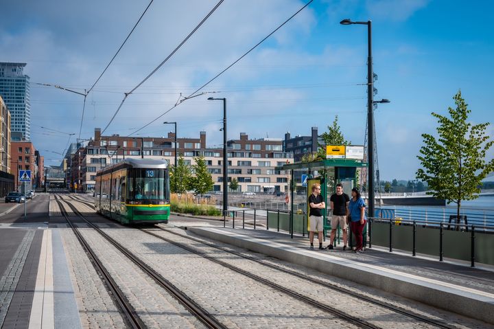 Kuvassa aurinkoisena kesäisenä päivänä raitiovaunu saapumassa pysäkille, jolla seisoo kaksi miestä ja nainen.