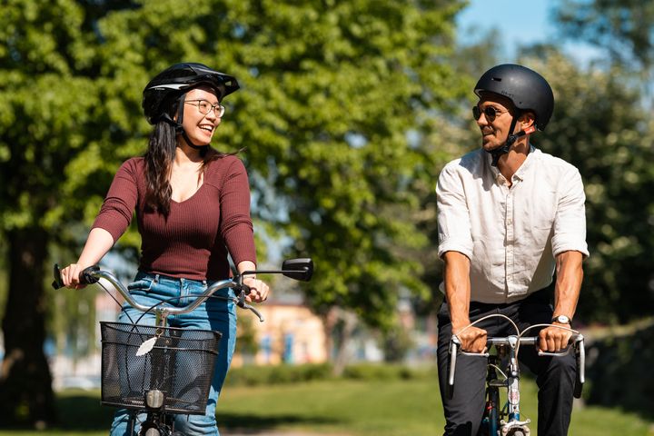 Två personer cyklar tillsammans i en park, båda bär cykelhjälmar och ler mot varandra.