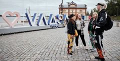 Four students stand and talk on a cobblestone street next to a large sign that reads "VAASA" with a heart symbol. A building is visible in the background.