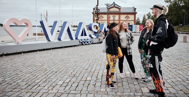 Four students stand and talk on a cobblestone street next to a large sign that reads "VAASA" with a heart symbol. A building is visible in the background.