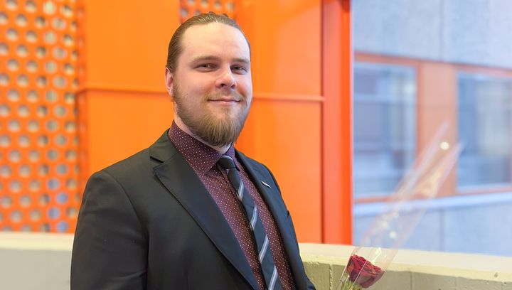 Freshly graduated Otto Mäkiniemi in formal attire holding a flower, standing in front of an orange wall at VAMK.