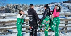 Four people wearing colorful winter outfits stand on a snowy wooden balcony, overlooking a scenic winter landscape with snow-covered trees and hills.