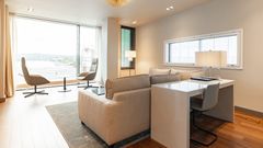 Kuva hotellihuoneesta, jossa on vaalea sohva, työpöytä ja kaksi nojatuolia ikkunan ääressä, josta avautuu näköala ulos.