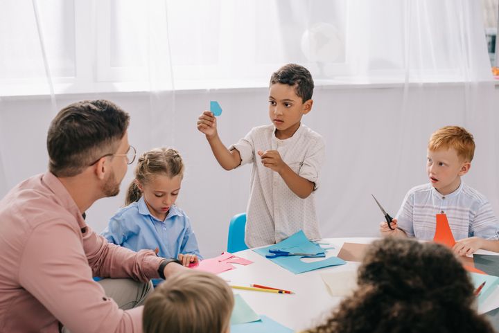 Kuvassa on lapsia ja miespuolinen opettaja pöydän ääressä. Pöydällä on askartelutarvikkeita. Yksi lapsista seisoo ja näyttää värikästä paperia opettajalle.