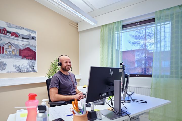 Mies istuu pöydän ääressä tietokoneen näytön edessä kuulokkeet päässään ja hymyilee.