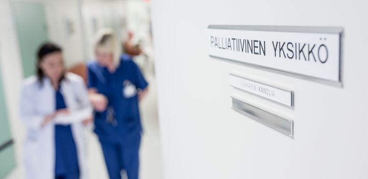 Kuvassa etualalla ovessa olevassa kyltissä lukee palliatiivinen yksikkö ja taustalla naislääkäri ja naishoitaja keskustelevat.