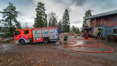 Pelastusharjoitus käynnissä: paloauto ja palomiehiä käyttämässä vesiletkuja rakennuksen edustalla.