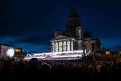 Kuvassa Helsingin Tuomiokirkko yötaivasta vasten. Kirkon portailla on valaistu lava, jossa soittajia rivissä. Portaiden alapäässä on paljon yleisöä.