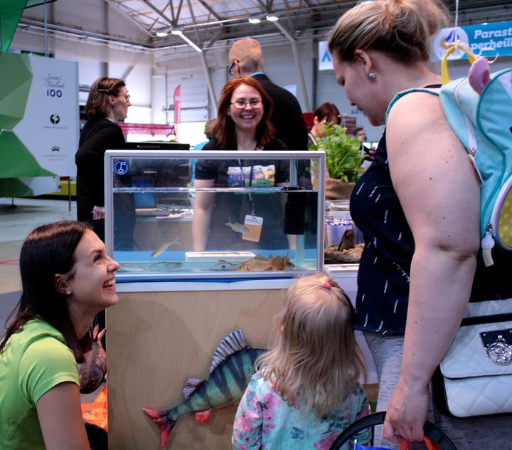 En grupp barn och vuxna tittar på en fisk i en liten tank vid en utställning.