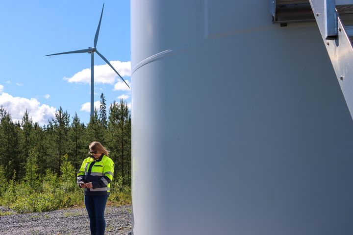 Person står vid en vindkraftverk i ett skogsområde.