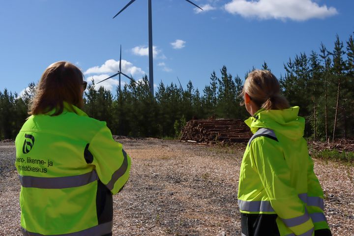 Kaksi henkilöä keltaisissa katselee tuulivoimaloita metsäisellä alueella.